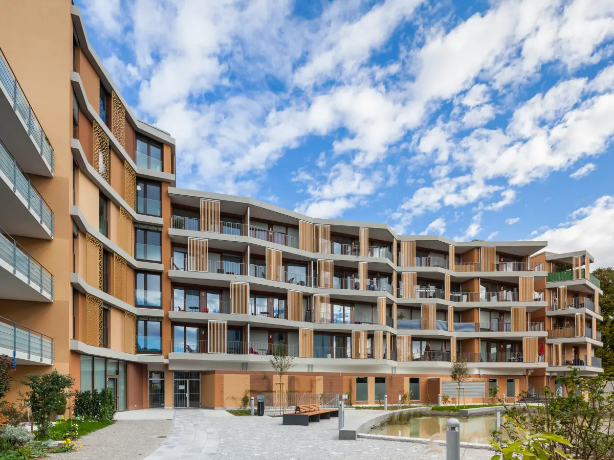 Moderno edificio residenziale con facciata curva, balconi aggettanti e cortile verde con vasca d'acqua.