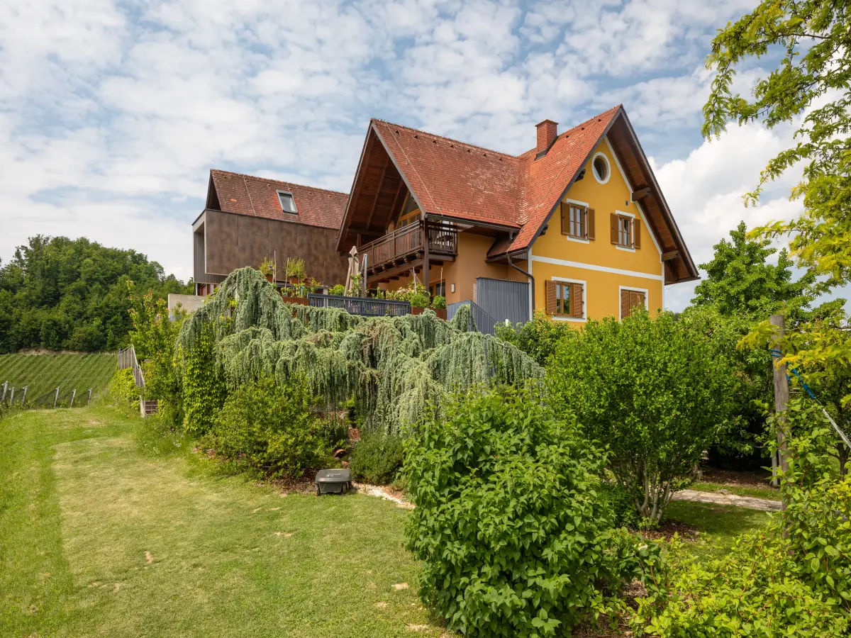 Gelbes Haus mit roten Dachziegeln und Balkon, umgeben von einem Garten und grüner Landschaft im Hintergrund.