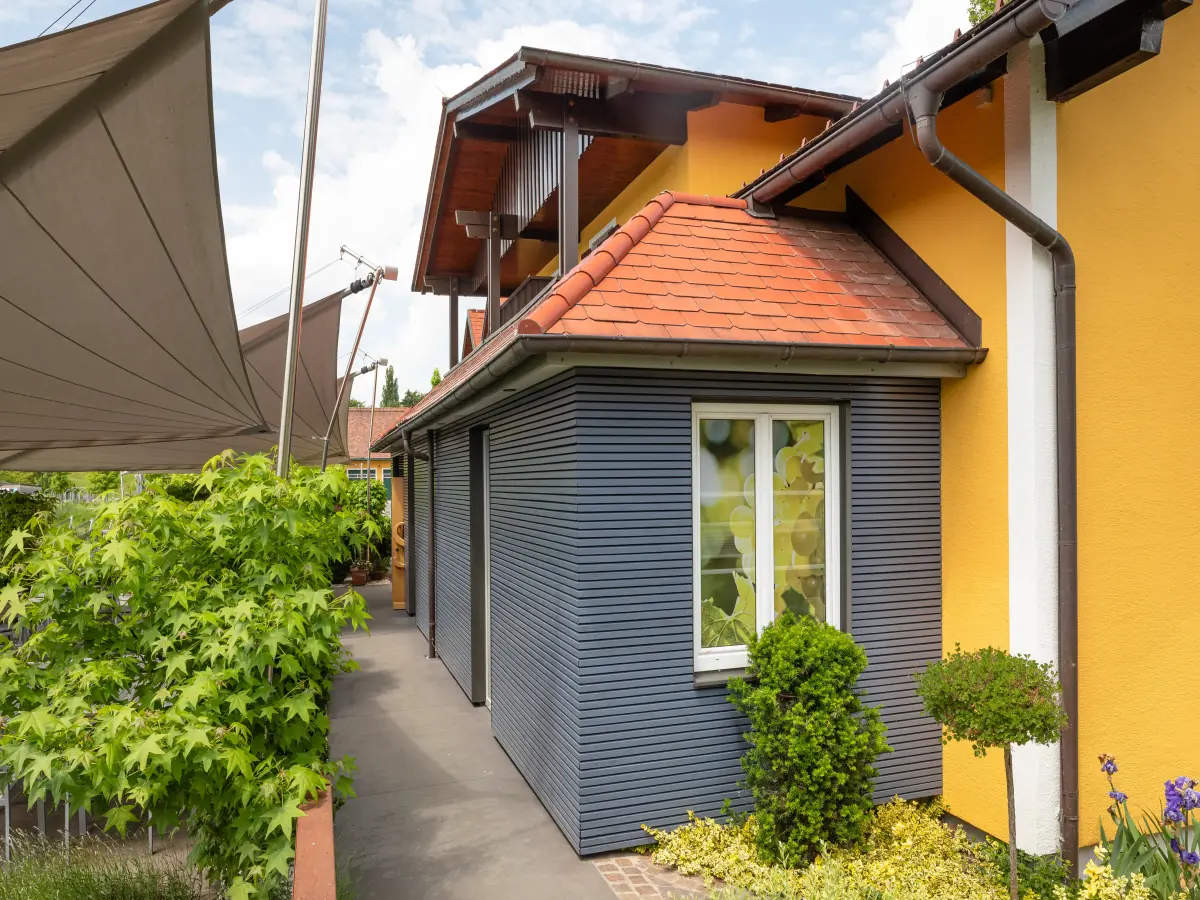 Modernes gelb-graues Haus mit Flachdach-Anbau, Pflanzen im Vordergrund und einer seitlichen Markise.
