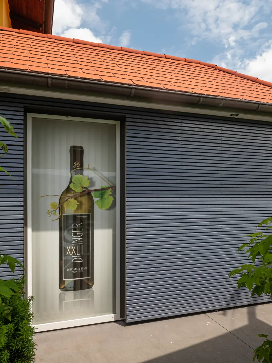 Detailansicht eines blaugrauen Hausanbaus mit Fenster, auf dem ein großes Werbemotiv für Weinflaschen abgebildet ist.