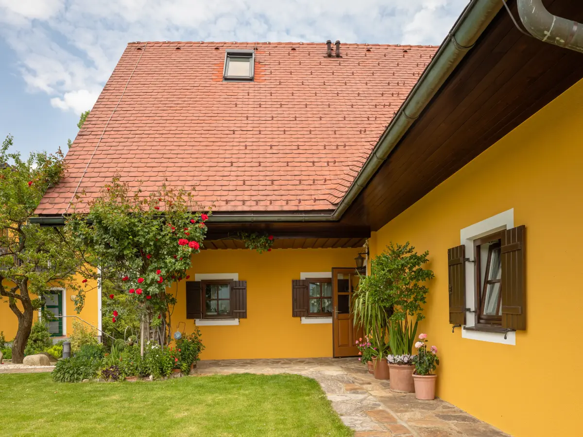 Gelbes Landhaus mit roten Dachziegeln, braunen Fensterläden, blühenden Pflanzen und gepflastertem Eingangsbereich.