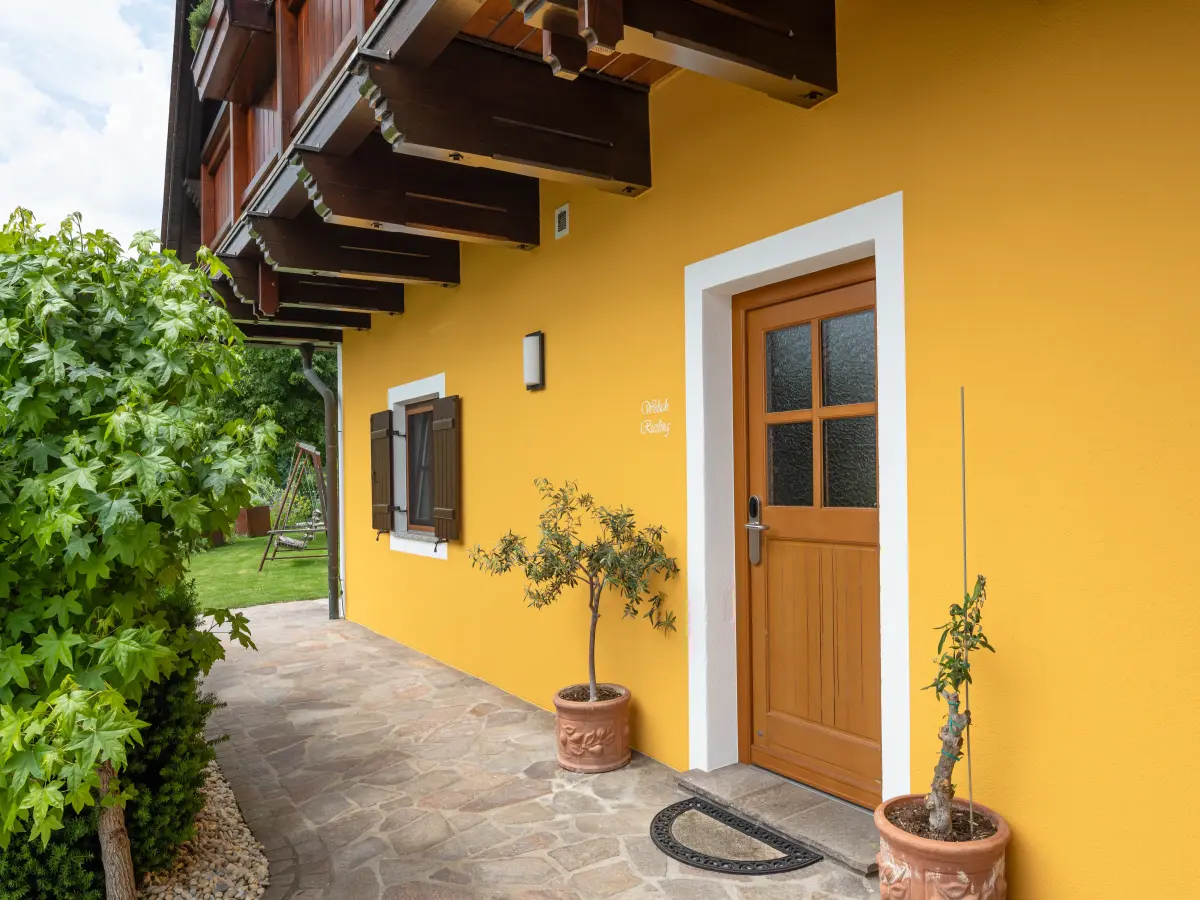 Gelbe Hausfassade mit Holztür, braunen Fensterläden, kleinen Olivenbäumen in Töpfen und gepflastertem Eingangsbereich.