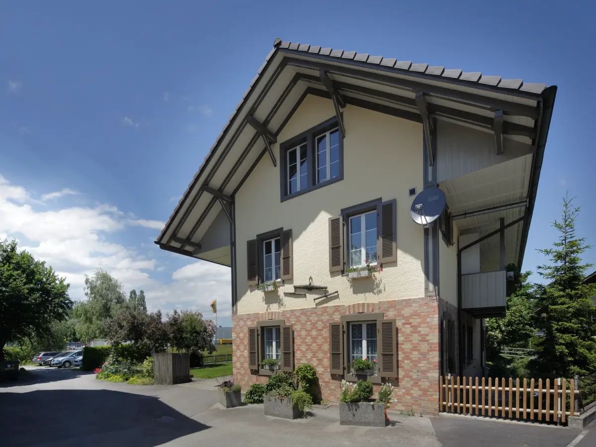 Traditionelles Haus mit cremefarbener Fassade, braunen Fensterläden und roten Klinkerelementen unter einem Satteldach.