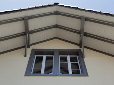 Nahaufnahme eines Giebels mit hellem Holzunterbau und grauen Balken. Darunter befindet sich ein graues Fenster mit weißen Sprossen, eingerahmt von einer cremefarbenen Fassade.