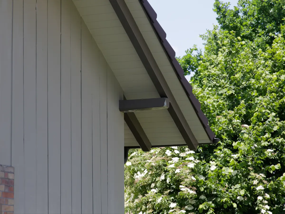 Nahaufnahme eines Giebels mit graubrauner Holzverschalung und Dachüberstand, umgeben von blühenden Bäumen im Hintergrund.