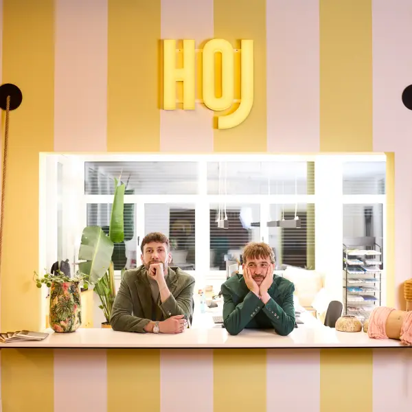Michael und Christof lehnen an einem Tresen und schauen in die Kamera. Die Wand dahinter ist gelb-gestreift und über den beiden hängt ein gelbes Neonschild mit der Aufschrift "HOJ" darauf.