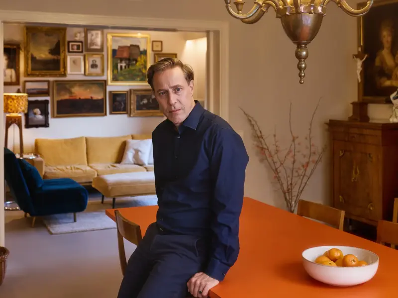 Jakob Bader sitzt lehnt an einem orangefarbenen Küchentisch und schaut ernst in die Kamera. Hinter ihm sind einige Gemälde an der Wand.