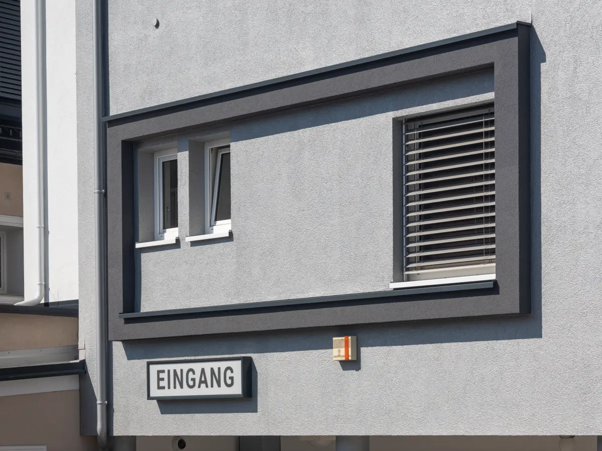 Detailansicht moderner Fassade mit grauem Fensterrahmen, Rollladen und "Eingang"-Schild.