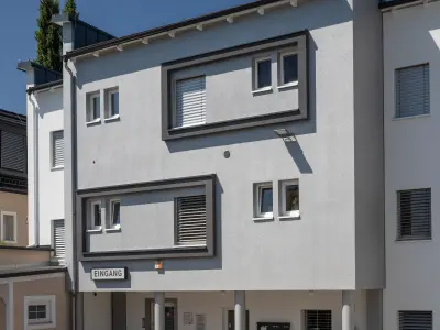 Die moderne Fassade ist weiß-grau mit rechteckigen Fenstern und Rollläden. Ein Schild mit „EINGANG“ deutet auf den Zugang hin. Die Architektur ist schlicht und geradlinig.