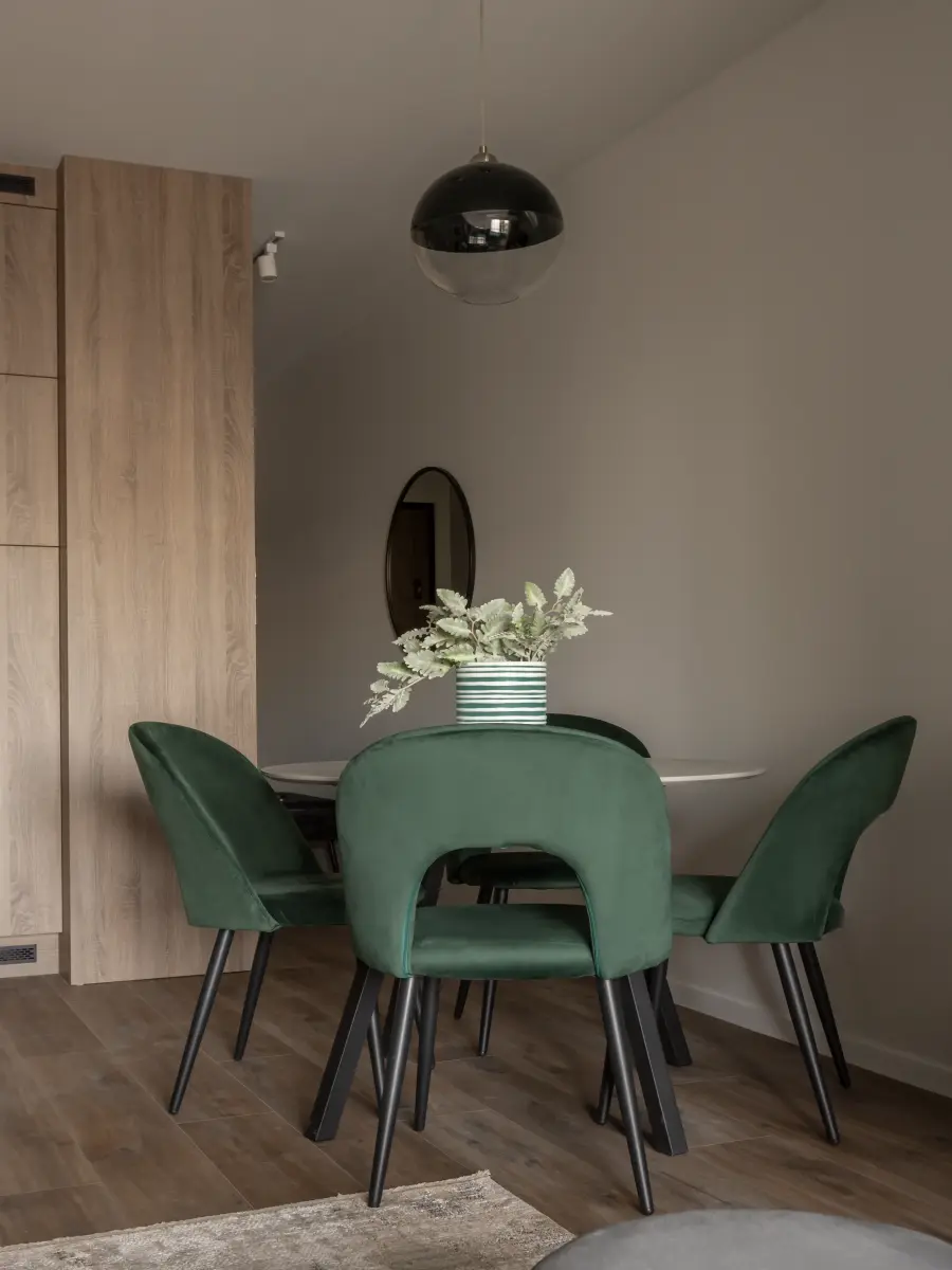 Grüne Samtstühle an einem weißen Esstisch, umgeben von Holzschränken und einer graubraunen Wand mit Spiegel.
