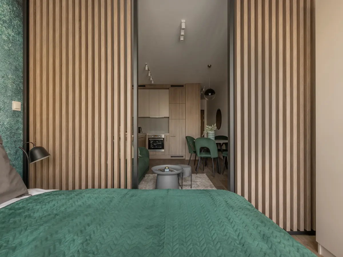 Blick von einem Bett mit grüner Steppdecke durch eine Öffnung mit Holzlamellen in einen offenen Küchen-/Essbereich.