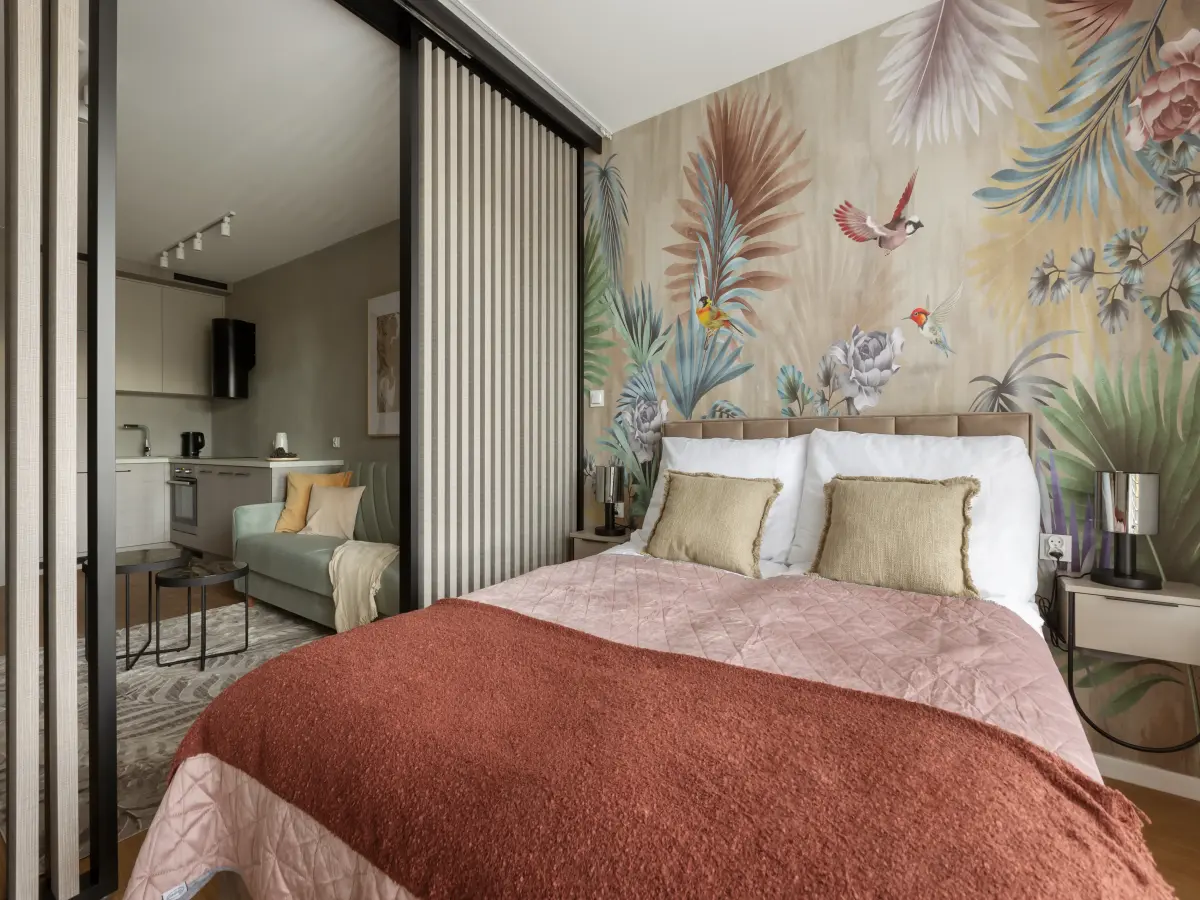 Bett mit rosa-rostfarbener Bettwäsche vor einer tropischen Dschungel-Tapete. Links ist der Wohnbereich durch eine Lamellentür sichtbar.