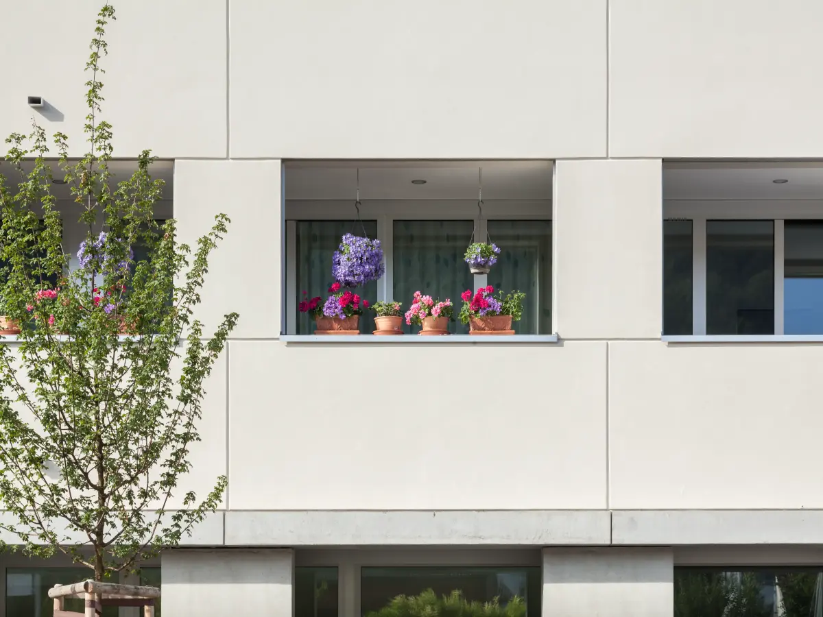 Detailansicht eines Balkons mit Blumen in Töpfen und Hängekörben, umgeben von einer modernen hellen Fassade.