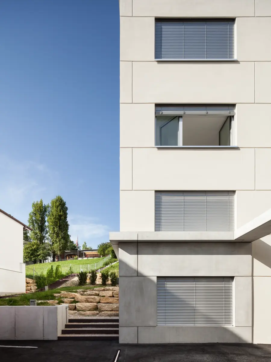 Frontale Ansicht eines modernen Gebäudes mit geschlossenen Rollläden, klaren Linien und einem kleinen Gartenbereich im Hintergrund.