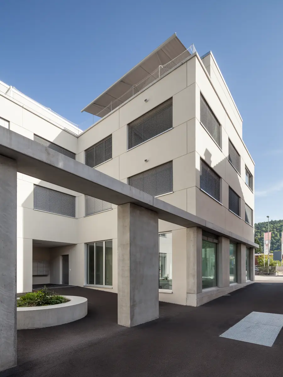 Modernes Gebäude mit klarer Architektur, großen Glasflächen im Erdgeschoss, Betonstützen und teilweise geschlossenen Rollläden, bei sonnigem Wetter.