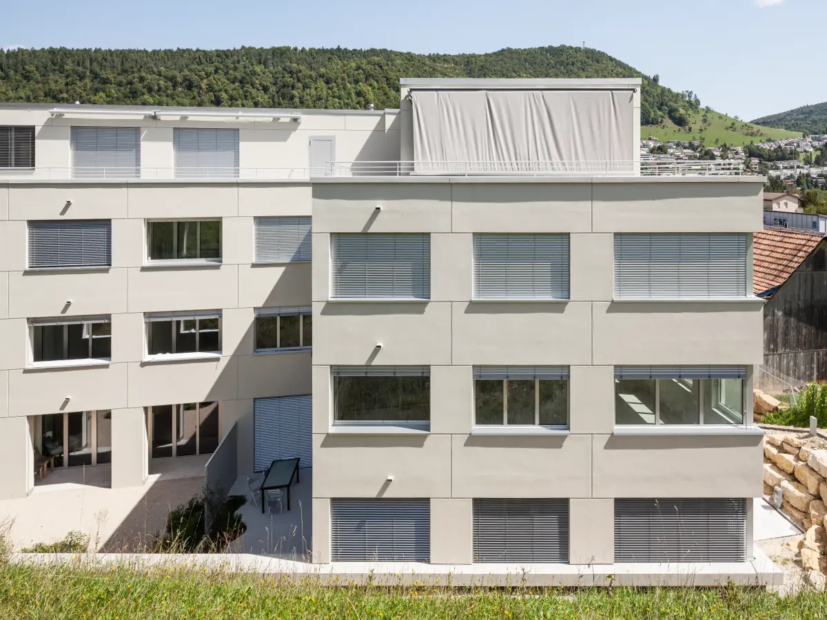 Modernes Wohngebäude mit hellgrauer Fassade, großen Fenstern und Rollläden, im Hintergrund eine grüne Hügelkulisse.