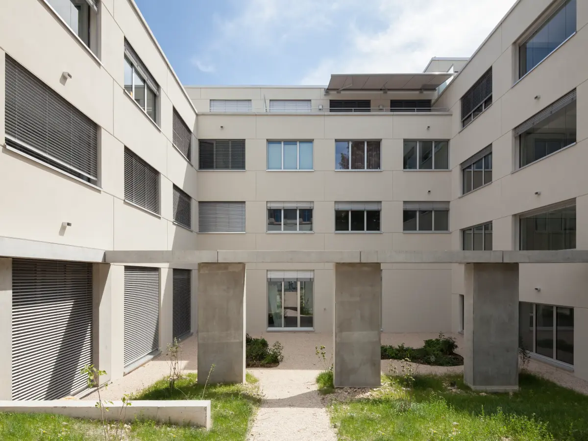 Innenhof eines modernen Wohngebäudes mit klarer Architektur, Betonstützen, Grünflächen und Kieswegen unter blauem Himmel.