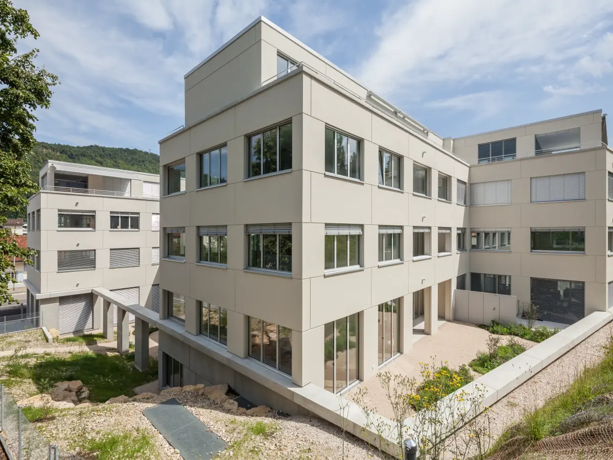 Außenansicht eines modernen Wohnkomplexes mit großen Fenstern, Kieswegen, Grünflächen und einer hügeligen Landschaft im Hintergrund.