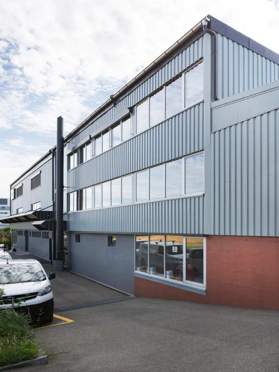 Seitenansicht eines Industriegebäudes der Merz Gips AG mit grauer Metallfassade, großen Fensterfronten und roter Basis, daneben geparkte Fahrzeuge unter einem leicht bewölkten Himmel.