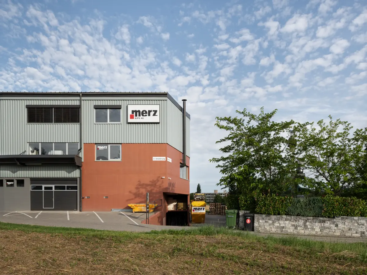 Industriegebäude der Firma Merz mit rot-grauer Fassade, Firmenlogo und Parkplatz, umgeben von Bäumen und leicht bewölktem Himmel.