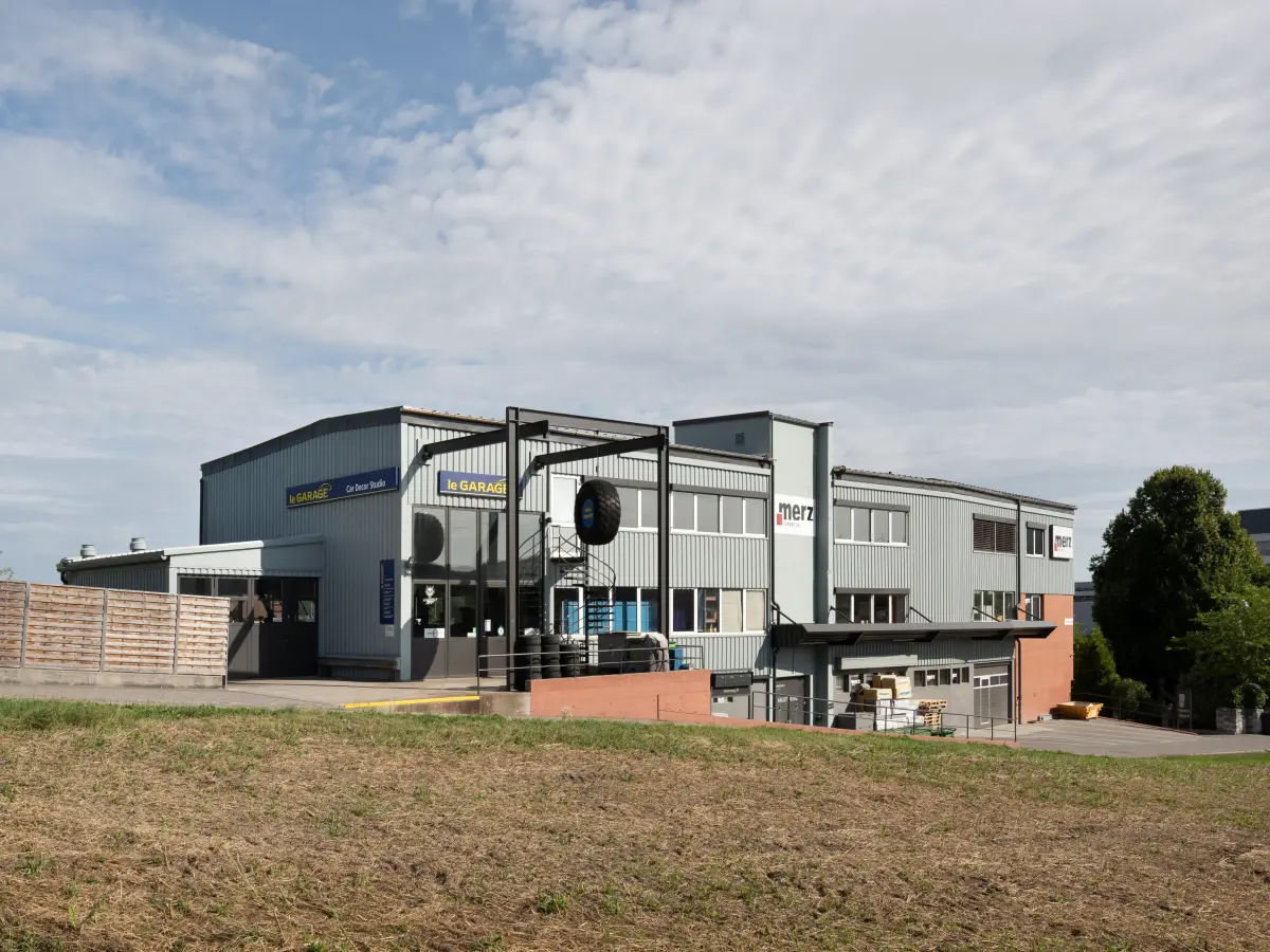 Industriegebäude mit der Aufschrift "Merz" und "le GARAGE", umgeben von einer ländlichen Landschaft und leicht bewölktem Himmel.