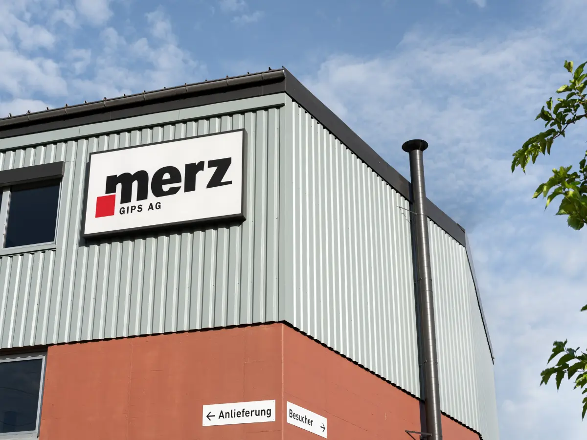 Detailaufnahme des Gebäudes der Merz Gips AG mit Firmenlogo, grauer Metallfassade und roter Unterkante, aufgenommen bei leicht bewölktem Himmel.