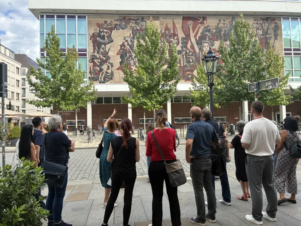 Eine Gruppe von Menschen steht auf dem Vorplatz des Kulturpalasts Dresden und betrachtet die große Wandmalerei an der Fassade.