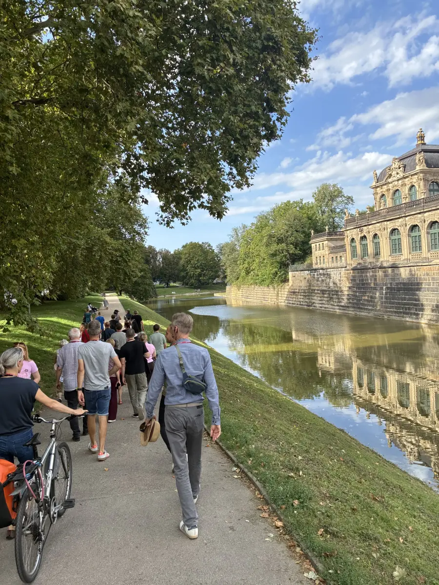 Eine Gruppe von Menschen geht am Wassergraben entlang, neben dem sich die begrünte Anlage und die barocke Fassade des Dresdner Zwingers spiegeln.
