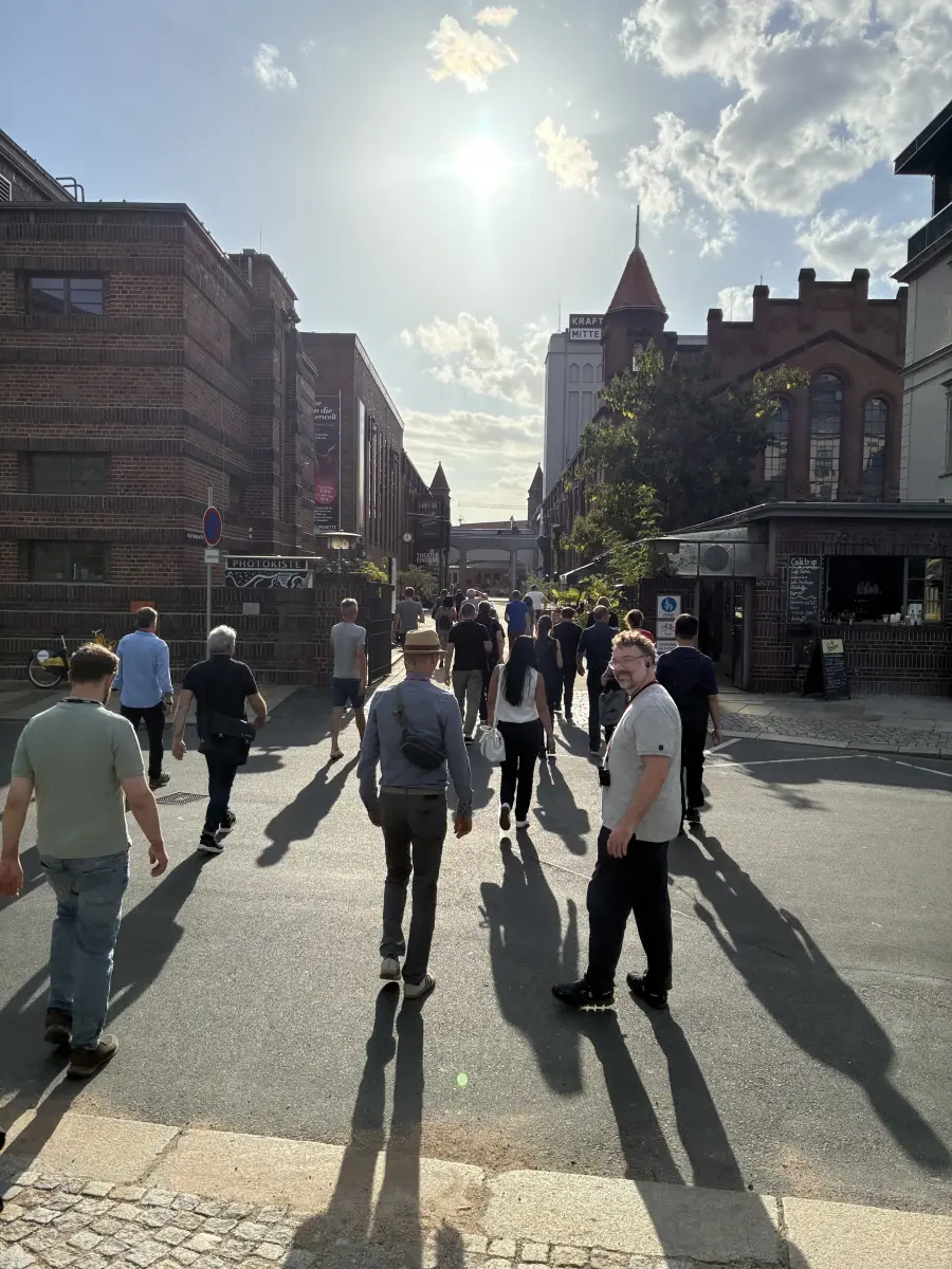 Menschenmenge auf einer Straße zwischen Backsteingebäuden auf dem Gelände des Kraftwerks Mitte; im Hintergrund ragt ein Turm mit rotem Kegeldach in den sonnigen Himmel.