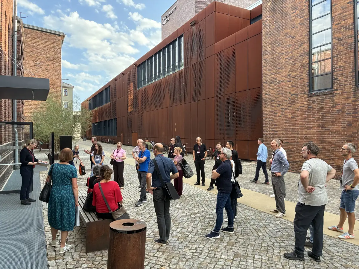 Eine Gruppe von Menschen nimmt an einer Führung im Innenhof zwischen Backsteinbauten und der modernen rostbraunen Fassade des Theaters Junge Generation teil.