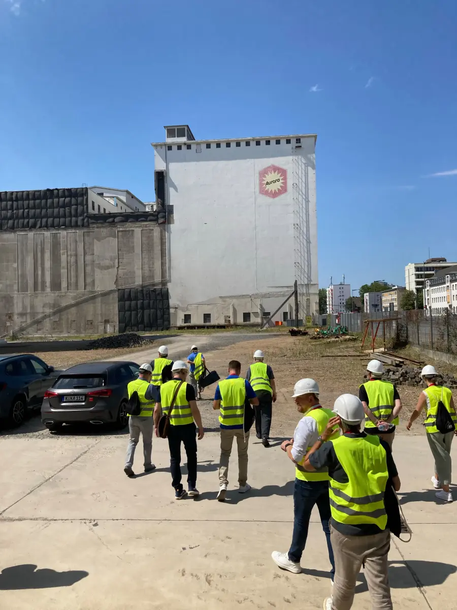 Gruppe mit Helmen und Warnwesten geht über ein Industriegelände vor großen Silos.