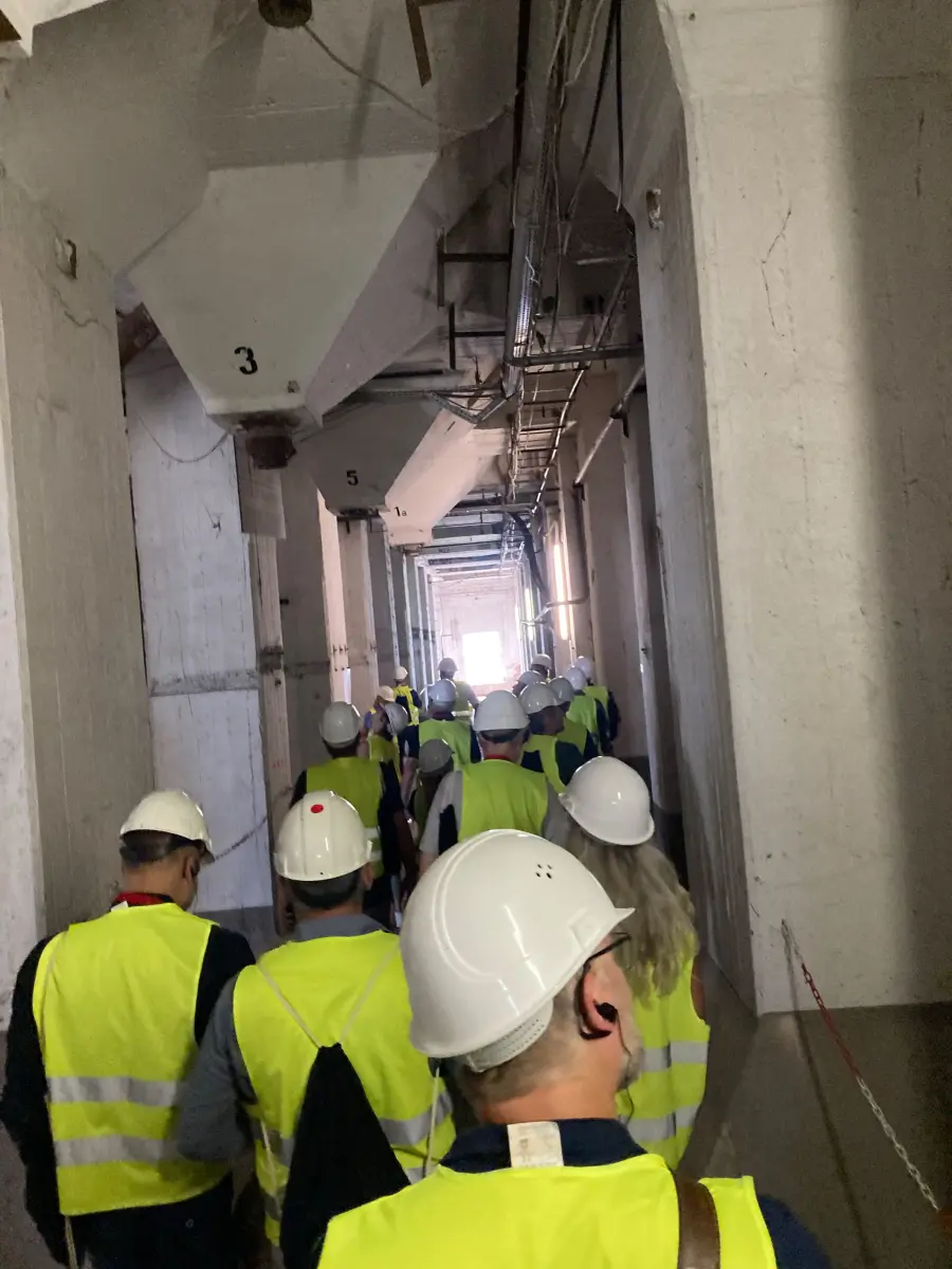 Gruppe von Personen mit Helmen und Warnwesten geht durch einen langen, industriellen Innenraum.