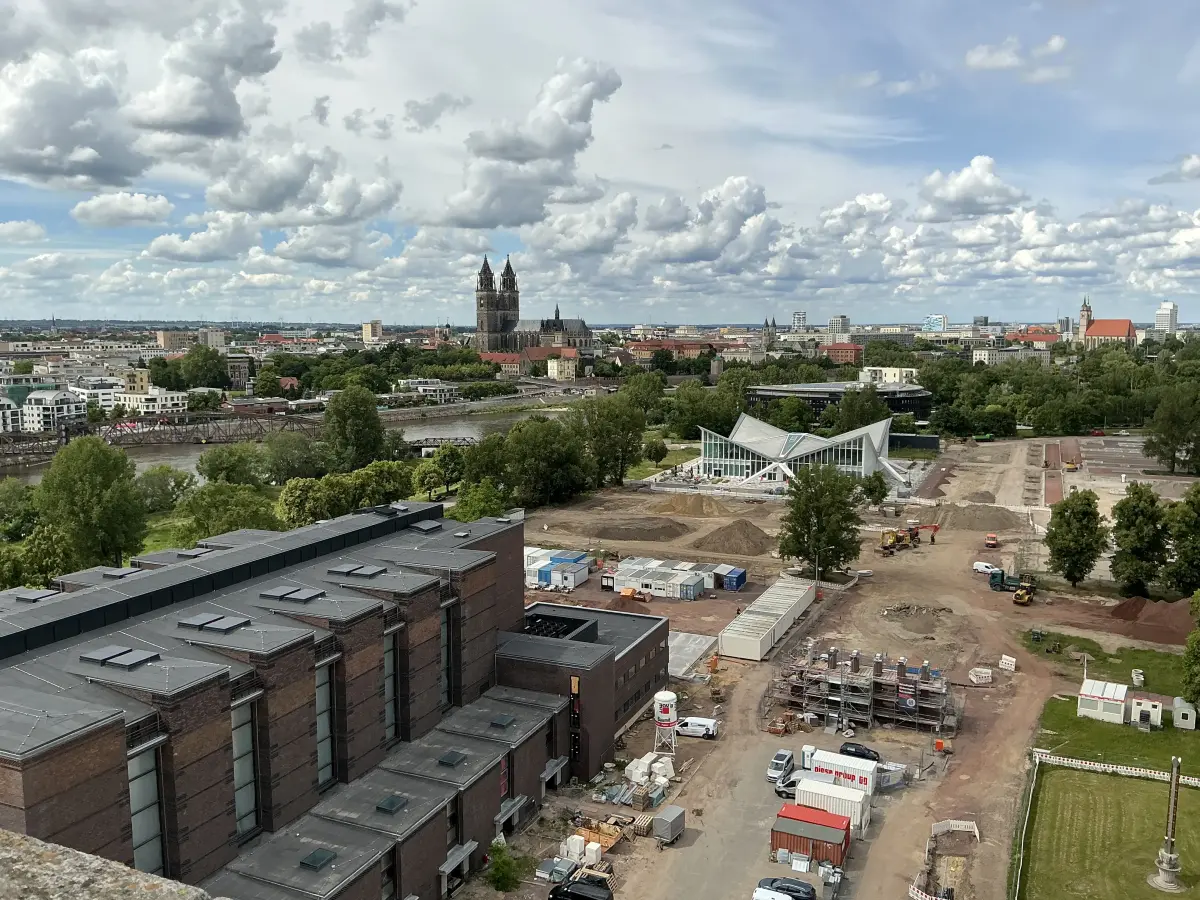 Stadtpanorama mit Fluss, Baustelle, weißem Gebäude mit gezacktem Dach und Backsteingebäude im Vordergrund.