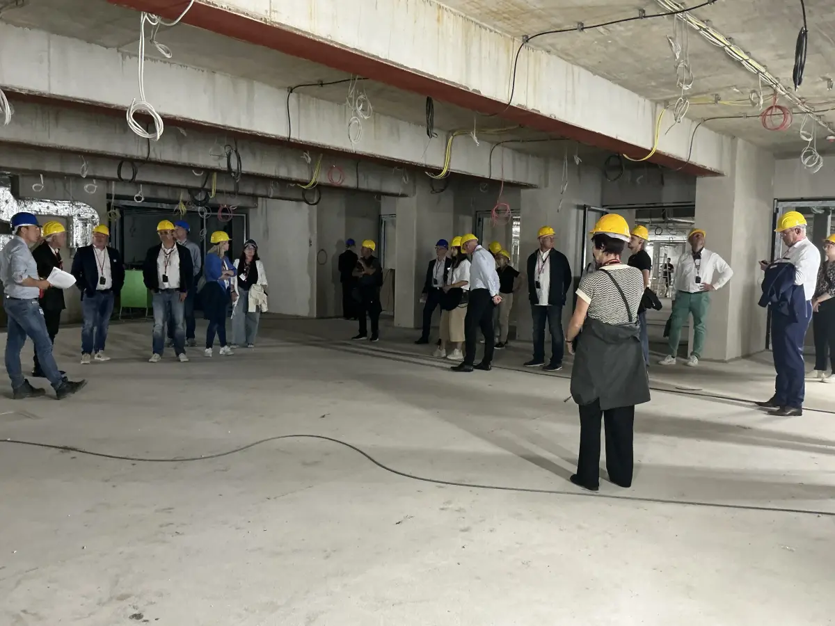 Rohbauetage mit Personen in gelben Schutzhelmen, Sichtbeton und freiliegenden Leitungen.