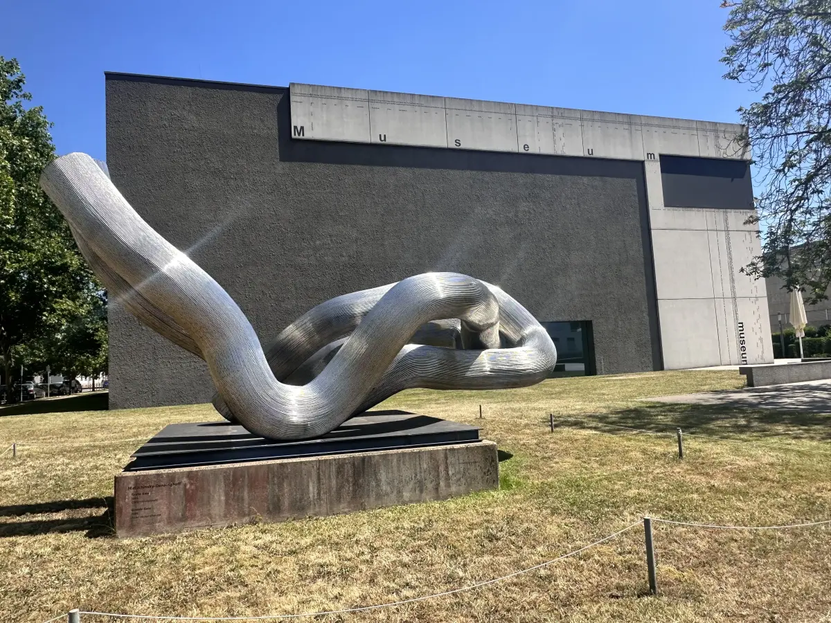 Silberne, geschwungene Metallskulptur auf einer Wiese vor einem modernen Museum mit dunkler Fassade und sonnigem Himmel.