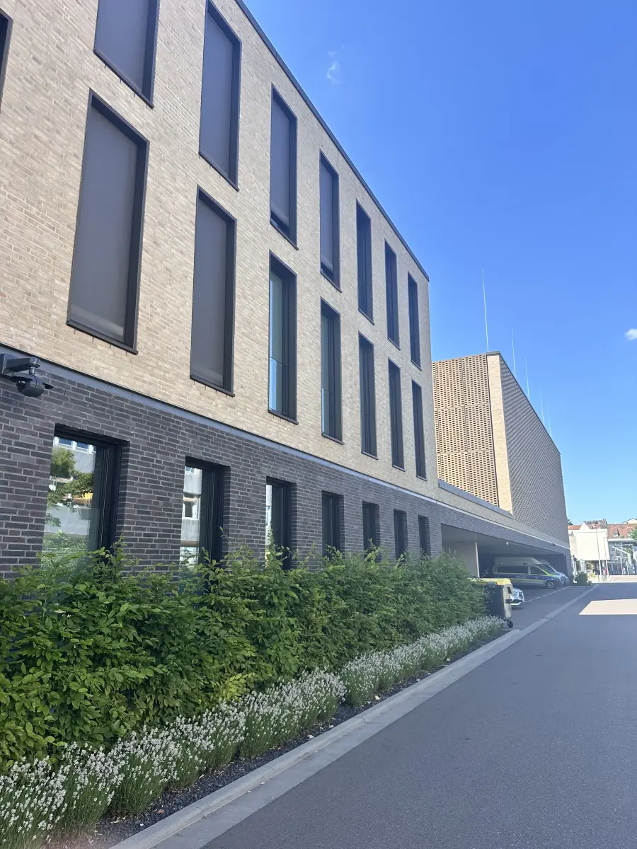 Seitliche Ansicht einer modernen Polizeiwache mit schmalen Fenstern, niedriger Bepflanzung und blauem Himmel.