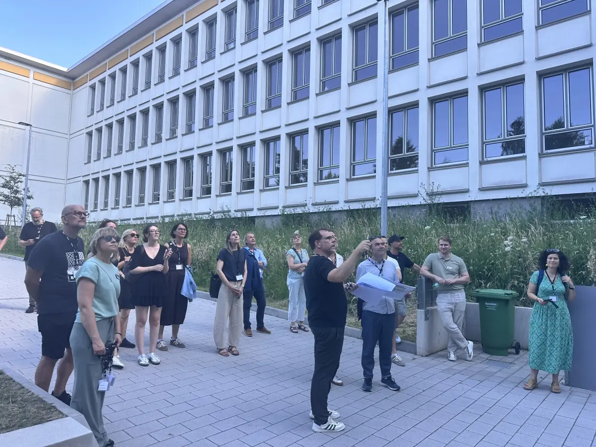 Gruppe von Personen steht auf dem Gehweg vor einem mehrstöckigen Gebäude, einige blicken nach oben und eine Person hält Papiere.