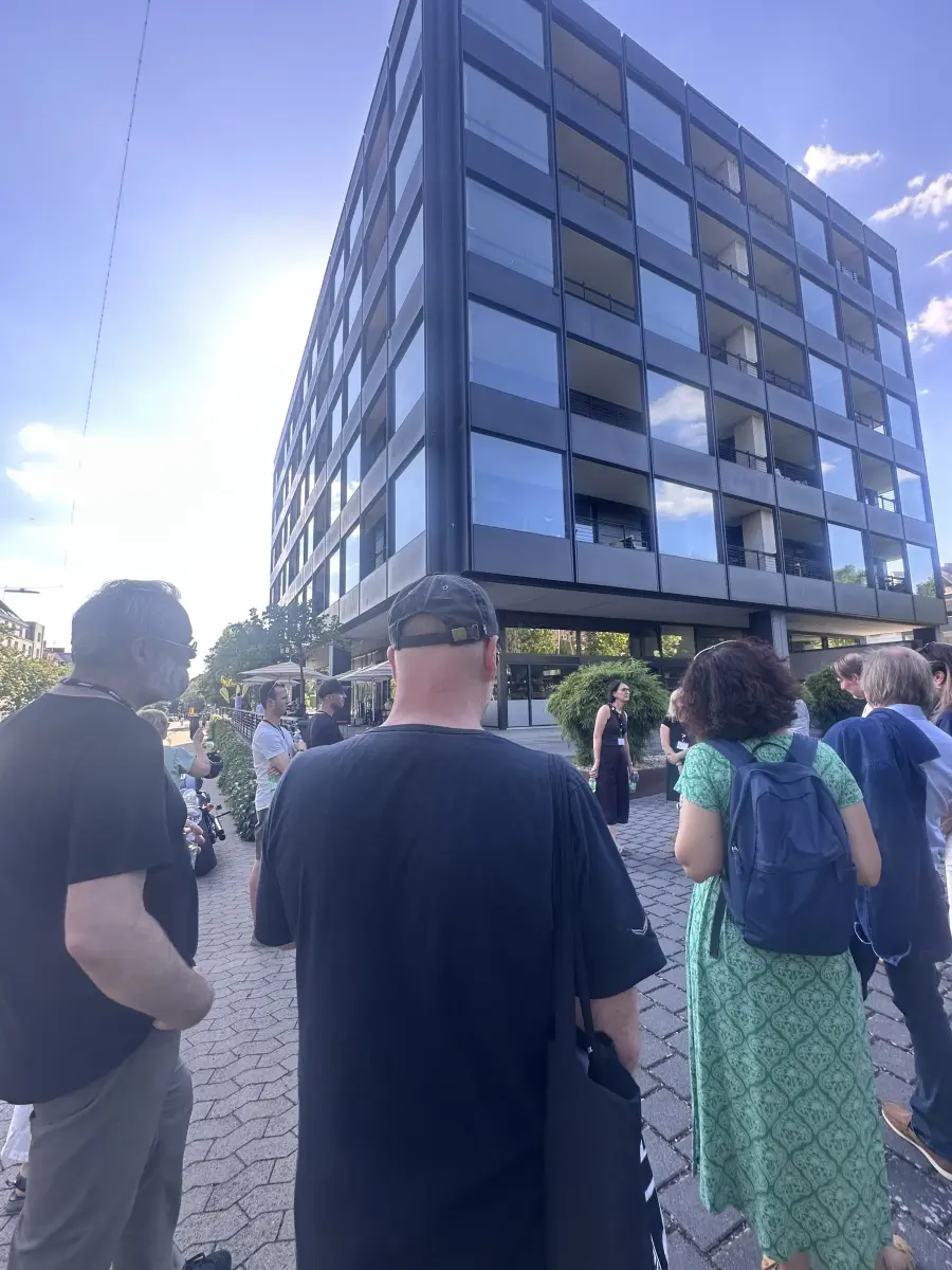 Gruppe von Menschen steht auf dem Bürgersteig vor einem modernen Glasgebäude bei sonnigem Himmel.