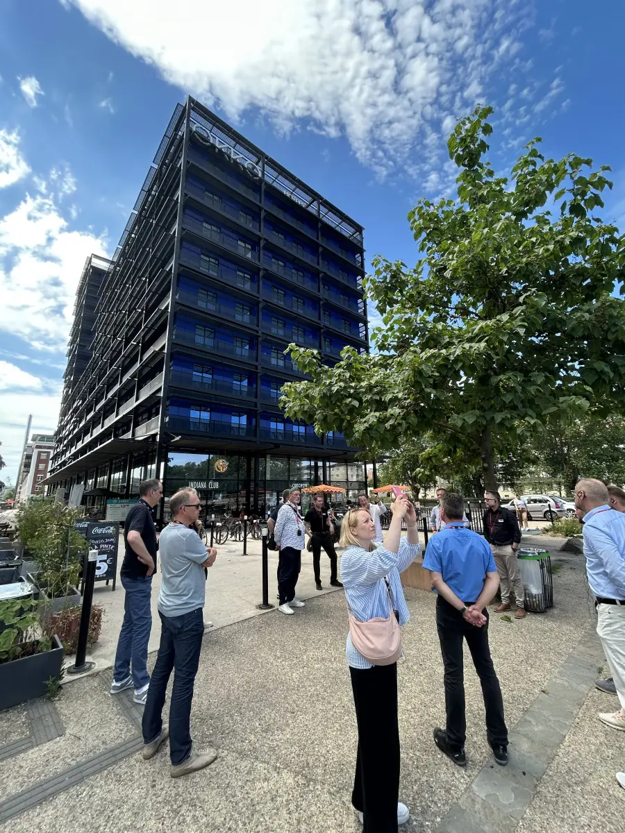  Menschen stehen vor einem modernen dunkelblauen Glasgebäude unter blauem Himmel.