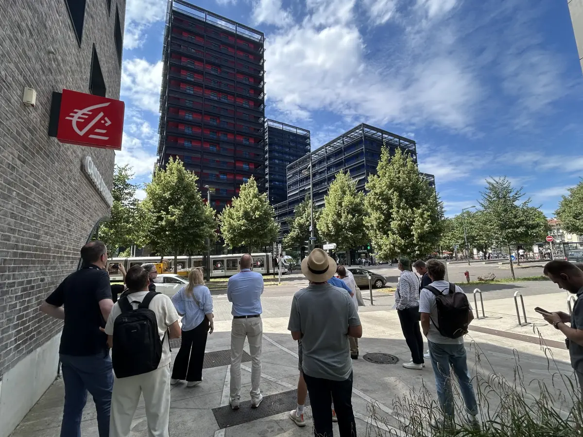 Gruppe von Menschen auf dem Gehweg vor modernen Glas‑ und Stahlhochhäusern mit roten Fassadenakzenten an einer städtischen Kreuzung.