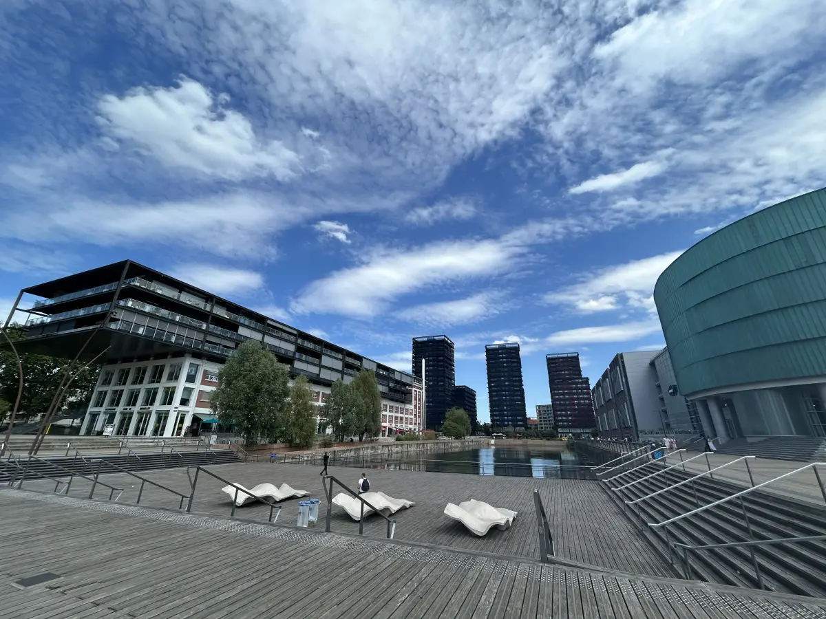 Uferpromenade mit Holzsteg und Liegen, spiegelndem Wasser und modernen Glas‑ und Stahlhochhäusern neben einem grünen Rundbau.