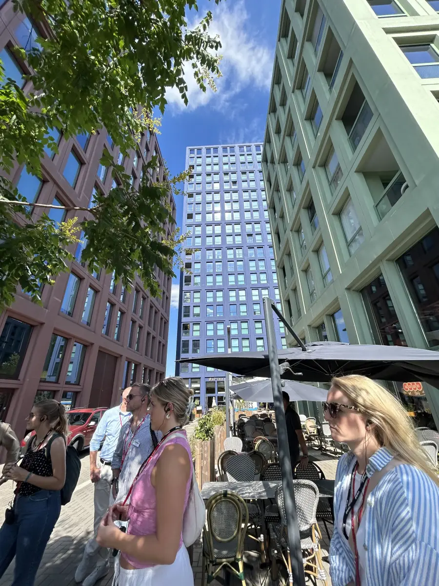 Tiefwinkelaufnahme von drei farbigen Kubusbauten mit regelmäßigen Fensterquadraten, Bäumen und Straßencafé im Vordergrund.