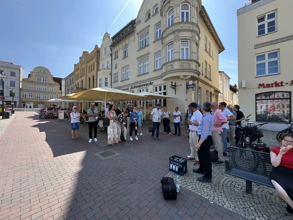 Gruppe von Menschen auf einem sonnigen Platz in der Altstadt vor historischen Gebäuden und einem Café mit Sonnenschirmen.