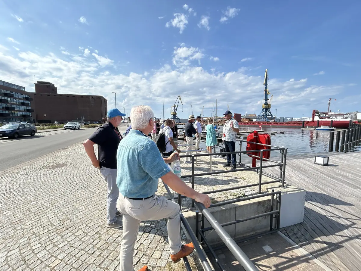 Gruppe von Menschen bei einem Spaziergang am Hafen; im Hintergrund Kräne, Wasser und Industrieanlagen unter blauem Himmel.