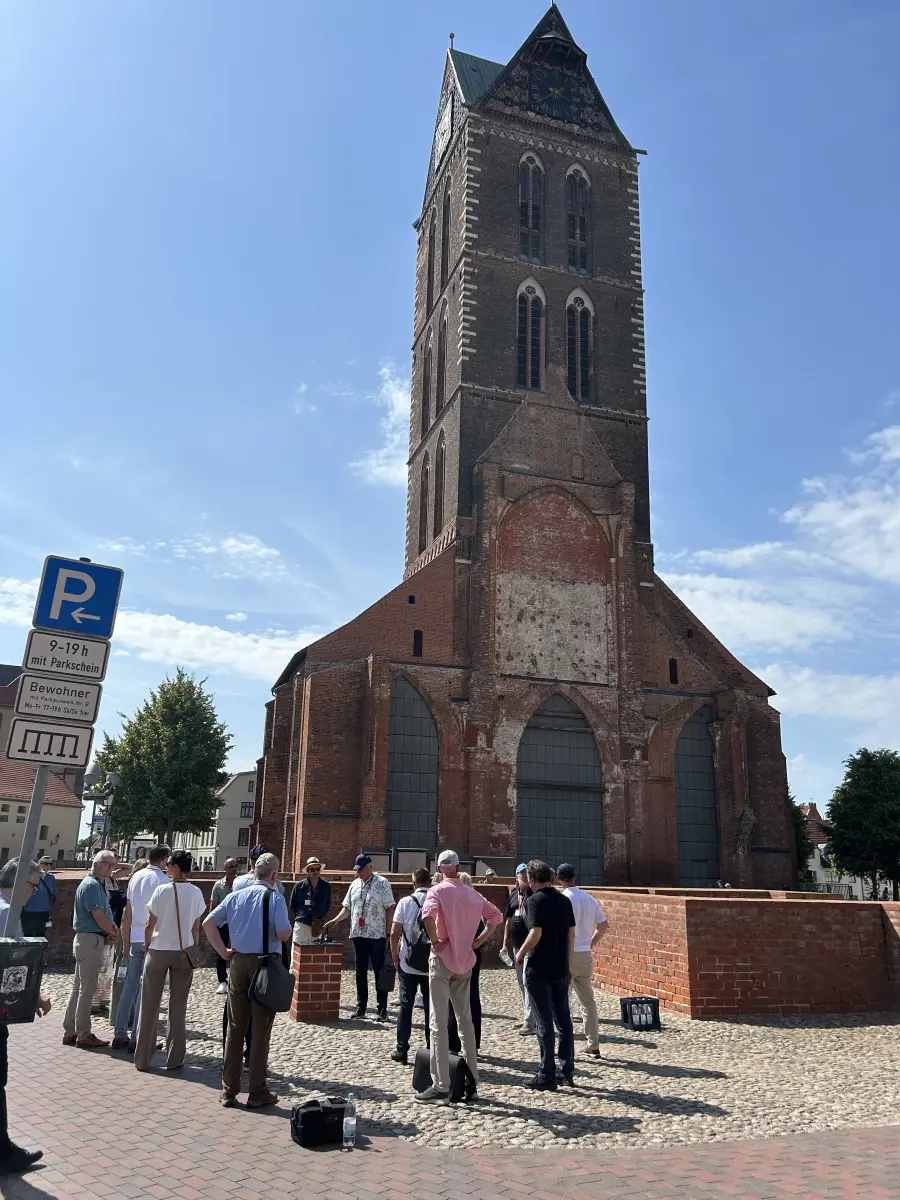 Gruppe von Menschen vor dem Turm einer historischen Backsteinkirche auf einem sonnigen Platz.