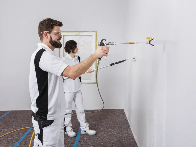 zwei Maler tragen mit Spritzgerät und einer Rolle Farbe auf eine Wand auf