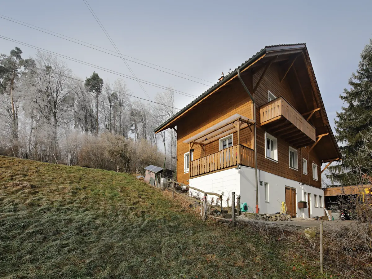 Ansicht eines traditionellen Holzhauses mit Balkon und Satteldach in ländlicher Umgebung an einem Hang, umgeben von herbstlichen Bäumen