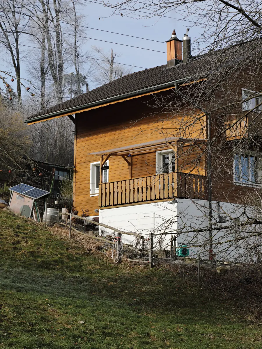 Seitenansicht eines Holzhauses mit Balkon und Schrägdach in ländlicher Umgebung.