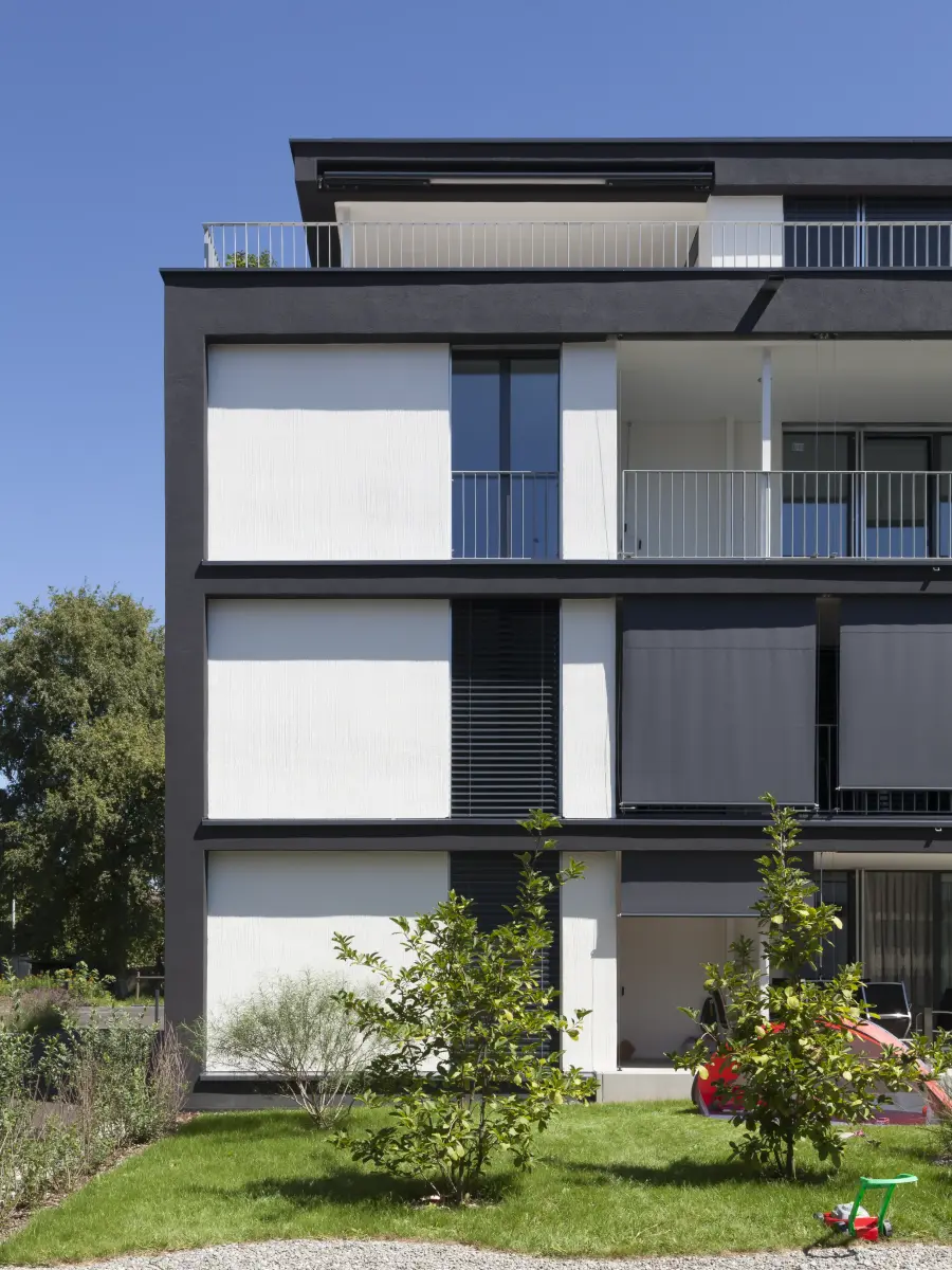 Detailansicht eines modernen Wohngebäudes mit weißer und dunkler Fassade, Balkonen und Schiebejalousien, umgeben von einer gepflegten Grünfläche.
