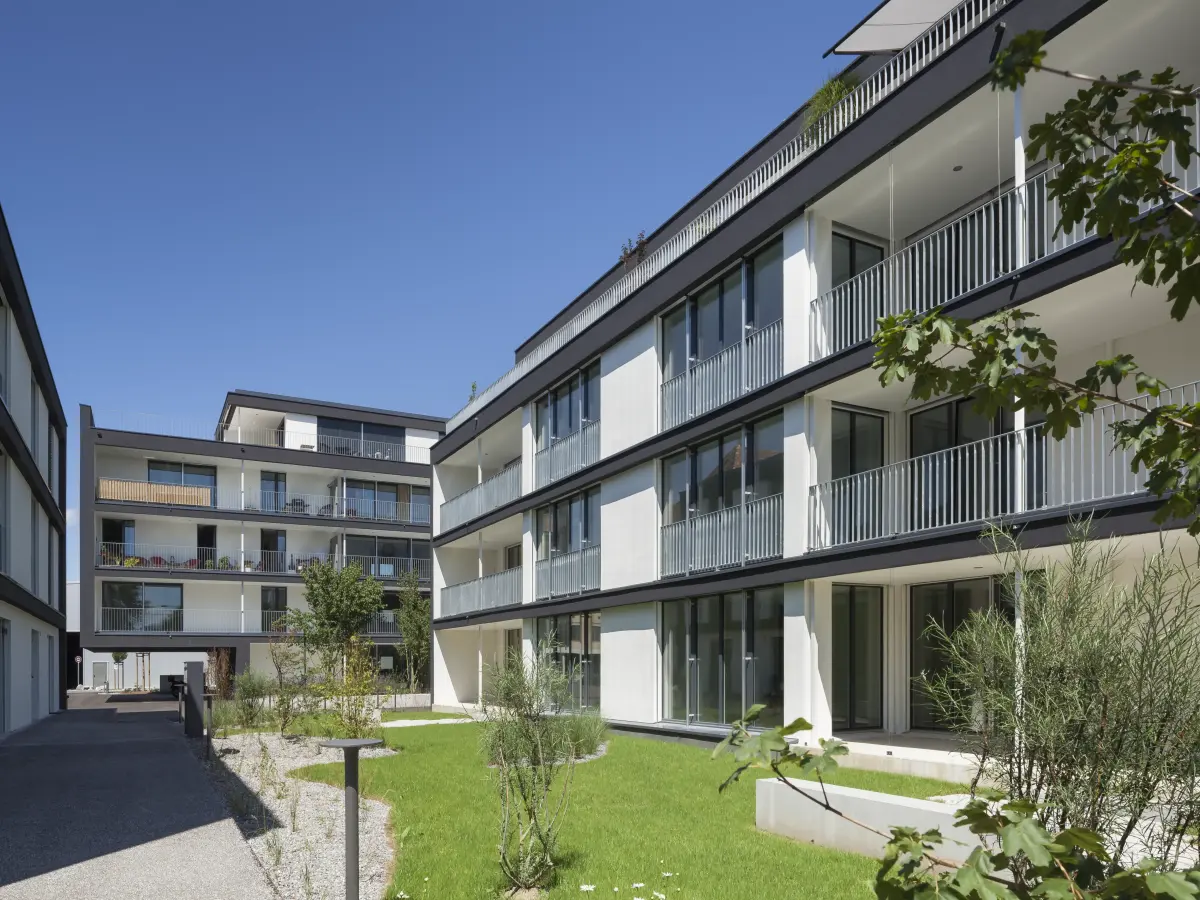 Innenhofansicht eines modernen Wohnkomplexes mit hellen Fassaden, großen Balkonen und einer gepflegten Grünfläche bei strahlend blauem Himmel.